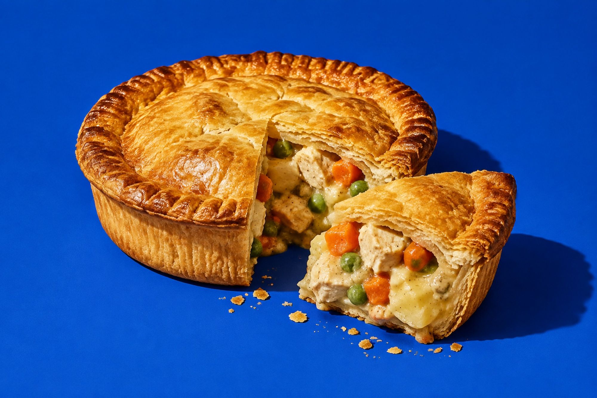 A golden chicken pie with a wedge cut out, showing chicken, carrot, peas and creamy gravy, on a cobalt blue backdrop